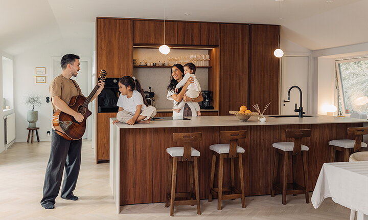 En familj i ett renoverat kök med varm träinredning. En vuxen spelar gitarr medan två barn och en vuxen sitter vid köksön.