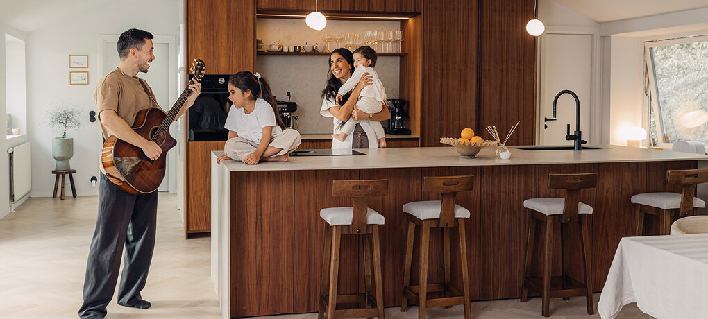 En familj i ett renoverat kök med varm träinredning. En vuxen spelar gitarr medan två barn och en vuxen sitter vid köksön.